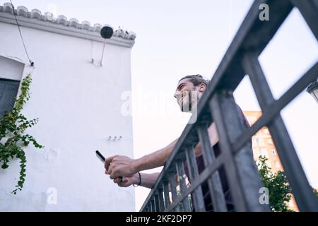 Der junge Kaukasusmann mit lang gesammeltem Haar lehnt sich glücklich auf ein schmutziges Geländer und schaut auf den Bildschirm seines Smartphones neben dem Haus Stockfoto