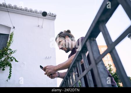 Junger kaukasischer Mann mit lang gesammeltem Haar, glücklich und auf einem schmutzigen Metallgeländer gestützt, der auf den Bildschirm seines Smartphones schaute Stockfoto