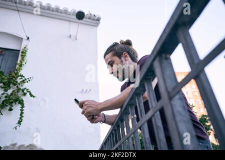 Der junge kaukasische Mann mit langen Haaren und gesammeltem Bart lehnte sich auf ein schmutziges Metallgeländer und schaute auf den Bildschirm seines Smartphones Stockfoto