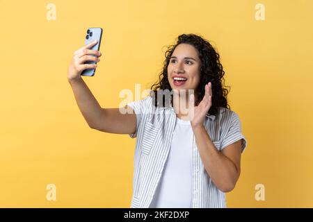 Porträt einer freundlichen Frau mit dunklen welligen Haaren, die das Smartphone hält, um Selfie zu machen oder einen Videoanruf zu tätigen, und mit der Hand die hallo-Geste winkt. Innenaufnahme des Studios isoliert auf gelbem Hintergrund. Stockfoto