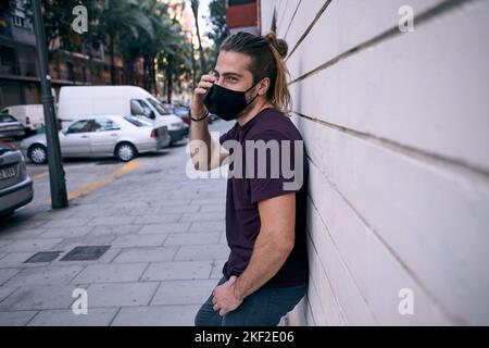 Kaukasischer junger Mann mit lang gesammeltem Haar, der mit der linken Hand in der Hosentasche steht und auf dem Smartphone mit einer Gesichtsmaske spricht Stockfoto