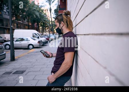 kaukasischer junger Mann mit lang gesammeltem Haar, der in einem schwarzen T-Shirt an einer Wand gelehnt auf den Smartphone-Bildschirm blickt, auf dem er ein schwarzes Gesicht trägt Stockfoto