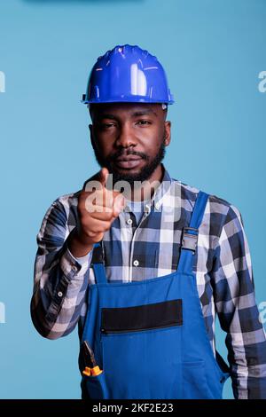 Porträt eines afrikanischen Mannes, der mit dem Zeigefinger auf die Kamera zeigt und auf blauem Hintergrund steht. Bauarbeiter im Fokus, die Richtung angeben und die Entscheidung treffen, Sie zu wählen, Studioaufnahme. Stockfoto