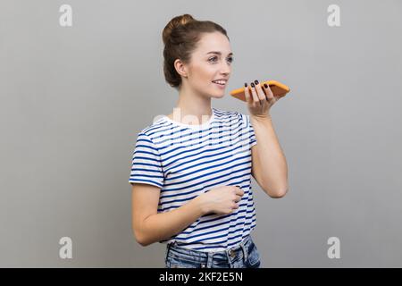 Intelligente Sprachtechnologie. Porträt einer Frau mit gestreiftem T-Shirt im Gespräch mit dem Handy mit virtuellen Assistenten, digitale Lautsprecher-Anwendung. Innenaufnahme des Studios isoliert auf grauem Hintergrund. Stockfoto