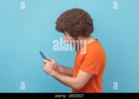 Seitenansicht eines staunenden Mannes mit Afro-Frisur, der ein orangefarbenes T-Shirt trägt und einen Beitrag über ein Mobiltelefon im sozialen Netzwerk liest und überrascht chattet. Innenaufnahme des Studios isoliert auf blauem Hintergrund. Stockfoto