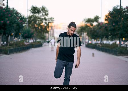 das schwarze T-Shirt des kaukasischen Mannes zerzauste lange Haare, ein Bart packte seinen rechten Fuß und seine Hand stand auf der Straße auf dem Stadtboulevard Stockfoto