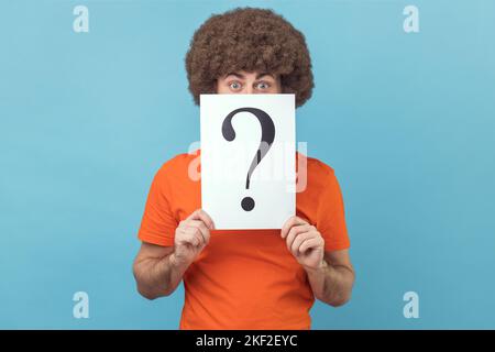 Mann mit Afro-Frisur in orangefarbenem T-Shirt, der die Hälfte des Gesichts hinter weißem Papier mit Fragezeichen versteckt, intelligente Lösung findet und um Rat bittet. Innenaufnahme des Studios isoliert auf blauem Hintergrund. Stockfoto