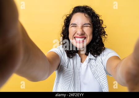 Porträt einer extrem glücklichen, schönen Frau mit dunkelwelligen Haaren, die ein Sichtbild macht, mit einem toothy Lächeln und geschlossenen Augen posiert, POV. Innenaufnahme des Studios isoliert auf gelbem Hintergrund. Stockfoto