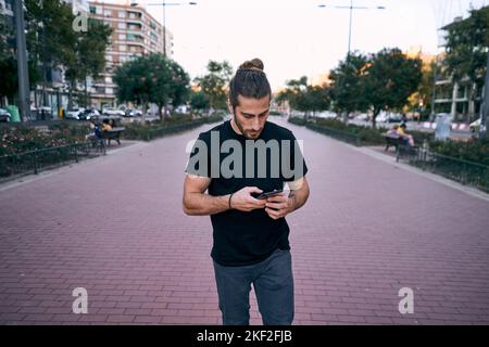 Kaukasischer junger Mann schwarzes T-Shirt lange Haare gesammelt Blick auf den Bildschirm seines smartphoe auf dem Boulevard in der Stadt Stockfoto