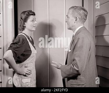 1930S SKEPTISCHE FRAU MIT SCHÜRZE AN DER EINGANGSTÜR IM GESPRÄCH MIT EINEM VERKÄUFER EIN REIFER MANN MIT GESCHÄFTSANZUG - S10189 HAR001 HARS HAUS LEBEN KOPIEREN RAUM HALBE LÄNGE DAMEN PERSONEN MÄNNER MITTLEREN ALTERS B & W MITTLEREN ALTERS MANN ZIELE HAUSFRAU ANZUG UND KRAWATTE VERKAUF HAUSFRAUEN ÄUSSERE GELEGENHEIT, HAUSFRAUEN BERUFE ZUSAMMENARBEIT MID-ADULT MID-ADULT FRAU VERKÄUFER SKEPTISCH SCHWARZ-WEISS KAUKASISCHEN ETHNIE HAR001 ALT MODISCH Stockfoto