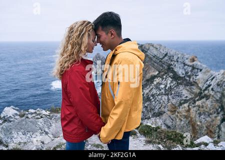 lateinischer Junge und blondes kaukasisches Mädchen im Regenmantel stehen im Profil gegenüber einander mit Stirn in Kontakt lächelnd die Hände vor dem halten Stockfoto