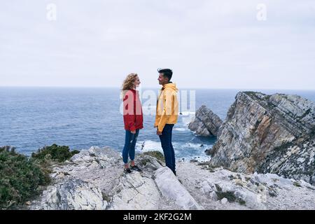 lateinischer Junge und kaukasisches Mädchen in roter Jacke und gelber Jeans stehen im Profil und sehen sich gegenseitig an und lächeln auf den großen Felsen Stockfoto