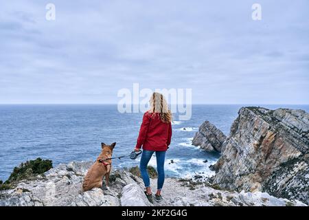 Junge kaukasische Frau mit einem großen blonden Hund an der Leine, der an einem bewölkten Tag, cabo de penas, die Meereswellen und den Horizont von den großen Felsen aus betrachtet Stockfoto