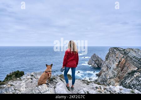 kaukasisches Mädchen mit einem großen Hund, der an einer Leine sitzt und an einem bewölkten Tag, cabo de, die Schönheit der Meereswellen und den Horizont der großen Felsen betrachtet Stockfoto