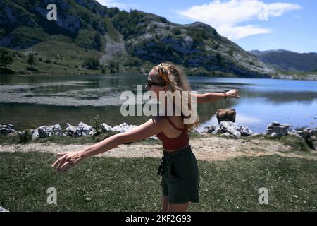 Blonde kaukasische Mädchen mit Sonnenbrille und Bogen im Haar Spaß in der Nähe des Sees Blick auf Kamera mit ausgestreckten Armen, covadonga asturias, spanien Stockfoto