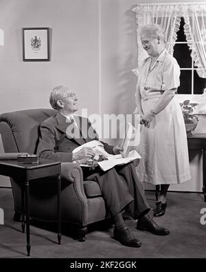 1940S 1950S ÄLTERER MANN SITZT IN EINEM SESSEL UND ÖFFNET EIN GESCHENK SEINER FRAU, DIE LÄCHELND VOR IHM STEHT - S3010 HAR001 HARS STARK ERFREUT FREUDE LIFESTYLE ZUFRIEDENHEIT FRAUEN VERHEIRATET STUDIO SHOT EHEPARTNER EHEMÄNNER ZU HAUSE LEBEN KOPIEREN RAUM IN VOLLER LÄNGE DAMEN PERSONEN FÜRSORGLICHE MÄNNER RUHESTAND ÄLTERER MANN ÄLTERER ERWACHSENER B&W PARTNER SENIOR FRAU RENTNER ANZUG UND KRAWATTE VOR GLÜCK FRÖHLICH HOHEN WINKEL SEINE FREIZEIT LÄCHELT VERBINDUNG KONZEPTUELLER JAHRESTAG EINFACH FRÖHLICH ÄLTERER MANN PERSÖNLICHER EIGENSINN ZUNEIGUNG ÄLTERE FRAU EMOTION IHN ZWEISAMKEIT FRAUEN SCHWARZ UND WEISS KAUKASISCHEN ETHNIZITÄT HAR001 ALTMODISCH Stockfoto