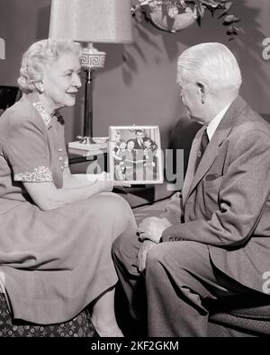 1950S SENIOREN IM WOHNZIMMER SITZEN UND AUF GERAHMTES FOTO IHRER KINDER UND ENKELKINDER SCHAUEN - S357 HAR001 HARS GROSSELTERN FAMILIEN FREUDE LIFESTYLE ÄLTERE FRAUEN VERHEIRATET GROSSELTERN STUDIO SHOT EHEPARTNER EHEMÄNNER ZU HAUSE LEBEN KOPIEREN RAUM FREUNDSCHAFT HALBE LÄNGE DAMEN PERSONEN INSPIRATION MÄNNER RUHESTAND ÄLTERER MANN ENKEL ÄLTERER ERWACHSENER B&W PARTNER ÄLTERER FRAU RENTNER GLÜCK ALTER OLDSTER UND STOLZ ZU GROSSMÜTTERN K ELDERS VERBINDUNG ENKELTOCHTER GROSSVÄTER GERAHMTER ENKEL ÄLTERER MANN ÄLTERE FRAU ZWEISAMKEIT FRAUEN SCHWARZ-WEISS KAUKASISCHE ETHNIE OMA OPA Stockfoto