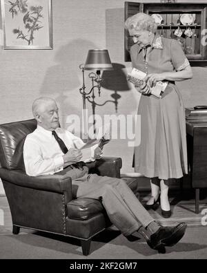1950S ÄLTERER MANN UND FRAU ZU HAUSE FRAU STEHEND MIT EINEM PAKET VON POST MANN SITZT AUF EINEM STUHL UND LIEST BRIEF - S390 HAR001 HARS ALTE MODE IM RUHESTAND 1 KOMMUNIKATION BALANCE TEAMWORK LIFESTYLE ÄLTERE FRAUEN VERHEIRATET EHEGATTEN EHEMÄNNER ZU HAUSE LEBEN KOPIE RAUM GANZKÖRPERDAMEN PERSONEN PFLEGE MÄNNER RUHESTAND SENIOR MANN SENIOR ADULT B & W PARTNER SENIOR FRAU RENTNER ALTER OLDSTERS LEBEN ZIMMER UND IN DER ÄLTEREN KONZEPTUELL STILVOLLES BUNDLE ÄLTERER MANN GRAUES HAAR ENTSPANNUNG ZWEISAMKEIT EHEFRAUEN ZU HAUSE SCHWARZ UND WEISSE KAUKASISCHE ETHNIE HAR001 ALTMODISCH Stockfoto