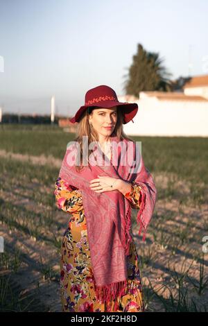 Stehende kaukasische reife Frau in rosa Schal geblümten Kleid und roten Hut lächelnd ruhig Blick auf die Kamera Stockfoto