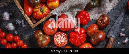 Viele verschiedene Arten, Formen und Größen von Tomaten in einer alten Holzkiste und auf einer dunklen Oberfläche, flach liegend, Draufsicht, Banner. Das Konzept der Ernte Stockfoto