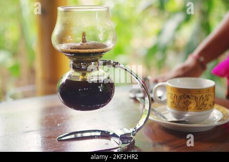 Eine Glas-Siphon-Kaffeemaschine; im Hintergrund eine Kaffeetasse mit einem traditionellen balinesischen Batik-Motiv auf einer Tee- und Kaffeeplantage in Ubud, Bali Stockfoto