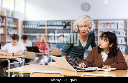 Ältere Lehrerin hilft Schülerinnen, die Gleichung in der Schulbibliothek zu lösen Stockfoto