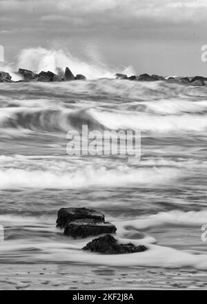 Eine Graustufen-Langzeitaufnahme von Wellen an einem Strand mit Felsen Stockfoto