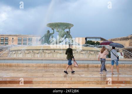 Valletta, Malta - 12. November 2022: Menschen, die an einem regnerischen Tag unter Sonnenschirmen am Triton-Brunnen spazieren Stockfoto