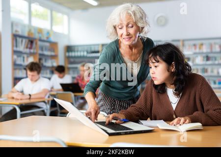 Ältere Lehrerin hilft Schülerinnen, die Gleichung in der Schulbibliothek zu lösen Stockfoto
