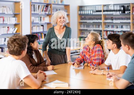 Ältere Lehrer, zusammen mit Jugendlichen Studenten, führt Tischklassen in der Schulbibliothek Stockfoto