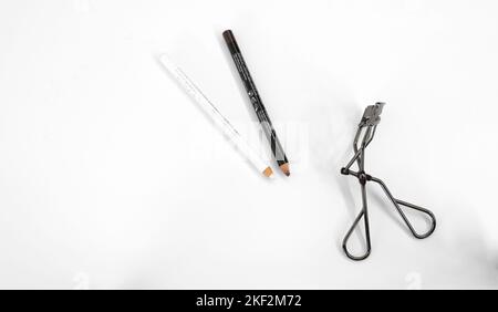 Brauner Lippenstift, weißer Eyeliner-Bleistift und Wimpern-Lockenstab auf weißem Hintergrund; Lippen- und Augen-Make-up und Kosmetik. Stockfoto