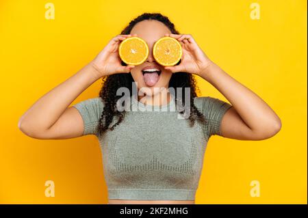 Fröhliche liebliche brasilianische oder latino junge lockige haarige Frau, in Sport-Outfits, stehend auf isoliertem orangefarbenem Hintergrund, hält zwei Hälften orange in den Händen neben den Augen, zeigt Zunge, lächelnd Stockfoto