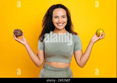 Hübsche, positive Mixed Race lockige, sportliche Frau, im Sportoutfit, hält frischen Apfel und süßen Donut, trifft die Wahl, steht auf isoliertem orangefarbenem Hintergrund, schaut fröhlich in die Kamera Stockfoto