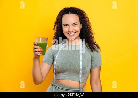 Gewichtsverlust, Entgiftung. Schöne brasilianische oder hispanische sportliche Frau, im Sport-Outfit und mit Zentimeter auf den Schultern, stehen auf isoliertem orangefarbenen Hintergrund, hält ein Glas frischen grünen Smoothie, lächelt Stockfoto