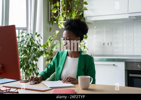 Fokussierte afroamerikanische Freiberuflerin sitzt am Tisch mit Computer und arbeitet zu Hause aus der Ferne. Stockfoto