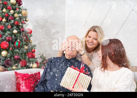 Enkelinnen schenken Großvater ein Geschenk. Glücklicher älterer Mann umarmt seine Enkelin, während er zu Weihnachten zu Hause ein Geschenk erhält. Stockfoto