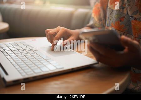 Nahaufnahme der Hand einer Frau mit dem Smartphone während der Arbeit am Laptop in einem Café. Stockfoto
