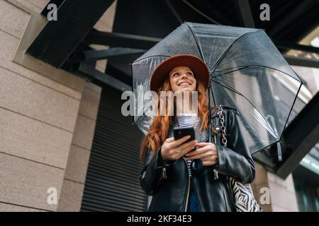 Low-Angle-Ansicht der positiven Ingwer junge Frau trägt eleganten Hut mit tippen Smartphone lächelnd Blick weg stehend mit transparentem Regenschirm Stockfoto