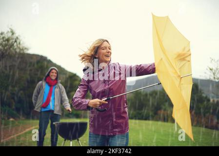 Gott sei Dank für meinen Regenschirm. Ein fröhliches Paar versucht, im Regen zu grillen, während sie einen Regenschirm lachend in der Hand hält. Stockfoto