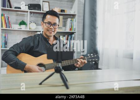 Asiatischer Musiklehrer Mann sendet live Gitarrenunterricht an Schüler online mit Handy. Asiatischer männlicher Musiker spielt Gitarre und singt live Video confer Stockfoto