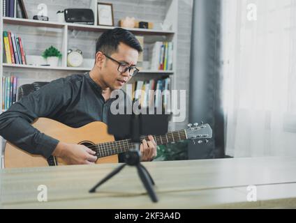 Asiatischer Musiklehrer Mann sendet live Gitarrenunterricht an Schüler online mit Handy. Asiatischer männlicher Musiker spielt Gitarre und singt live Video confer Stockfoto