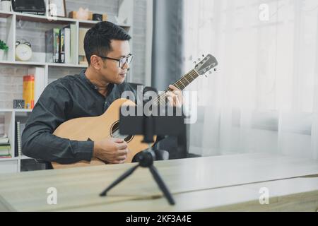 Asiatischer Musiklehrer Mann sendet live Gitarrenunterricht an Schüler online mit Handy. Asiatischer männlicher Musiker spielt Gitarre und singt live Video confer Stockfoto