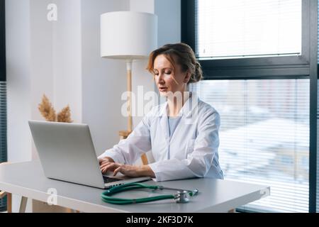 Porträt einer blonden Praktizierenden in weißem Mantel, die am Laptop-Computer beim Tippen arbeitete und auf dem Bildschirm aussutierte, die bei Licht am Tisch saß Stockfoto