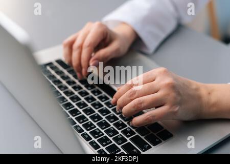 Nahaufnahme einer unkenntlich denkenden Ärztin in weißer medizinischer Uniform mit Laptop-Tastatur, die im modernen Büro am Schreibtisch sitzt. Stockfoto