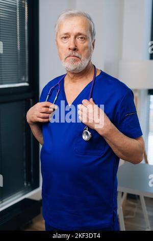 Vertikales Porträt eines traurigen überarbeiteten älteren männlichen Arztes in dunkelblauer medizinischer Uniform, der im Krankenhausbüro steht und die Kamera anschaut. Stockfoto