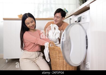 Junge Asiaten heirateten glückliches Paar lächelnd und machen Wäsche zu Hause. Freund und Freundin, die ihre Kleidung vor dem Laden der Waschmaschine zusammenlegen Stockfoto
