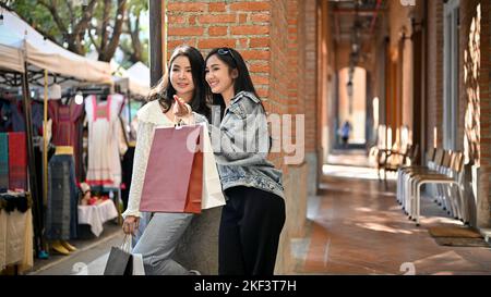 Zwei charmante junge asiatische Reisende mit ihren Einkaufstaschen gehen gemeinsam gerne Thai-lokale Produkte an der Einkaufsstraße ein. Stockfoto