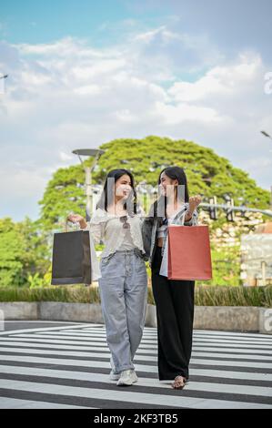 Porträt, schöne und fröhliche junge asiatische Freundinnen mit ihren Einkaufstaschen, überqueren den Fußgängerüberweg, genießen den Einkauf in der Einkaufsstraße toge Stockfoto