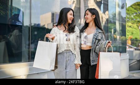 Zwei attraktive und fröhliche junge asiatische Frauen mit ihren Einkaufstüten reden und lachen beim Spaziergang entlang des Einkaufszentrums draußen Stockfoto