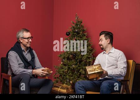 Älterer Mann und sein erwachsener Sohn lächeln und öffnen Weihnachtsgeschenke auf rotem Wandhintergrund und grünem Weihnachtsbaum Stockfoto